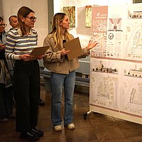 Die beiden Architekturstudentinnen stehen links neben ihren Planungsskizzen und erklären den Inhalt. Das Jurymitglied Peter Lesko steht rechts im Bild und hört zu.