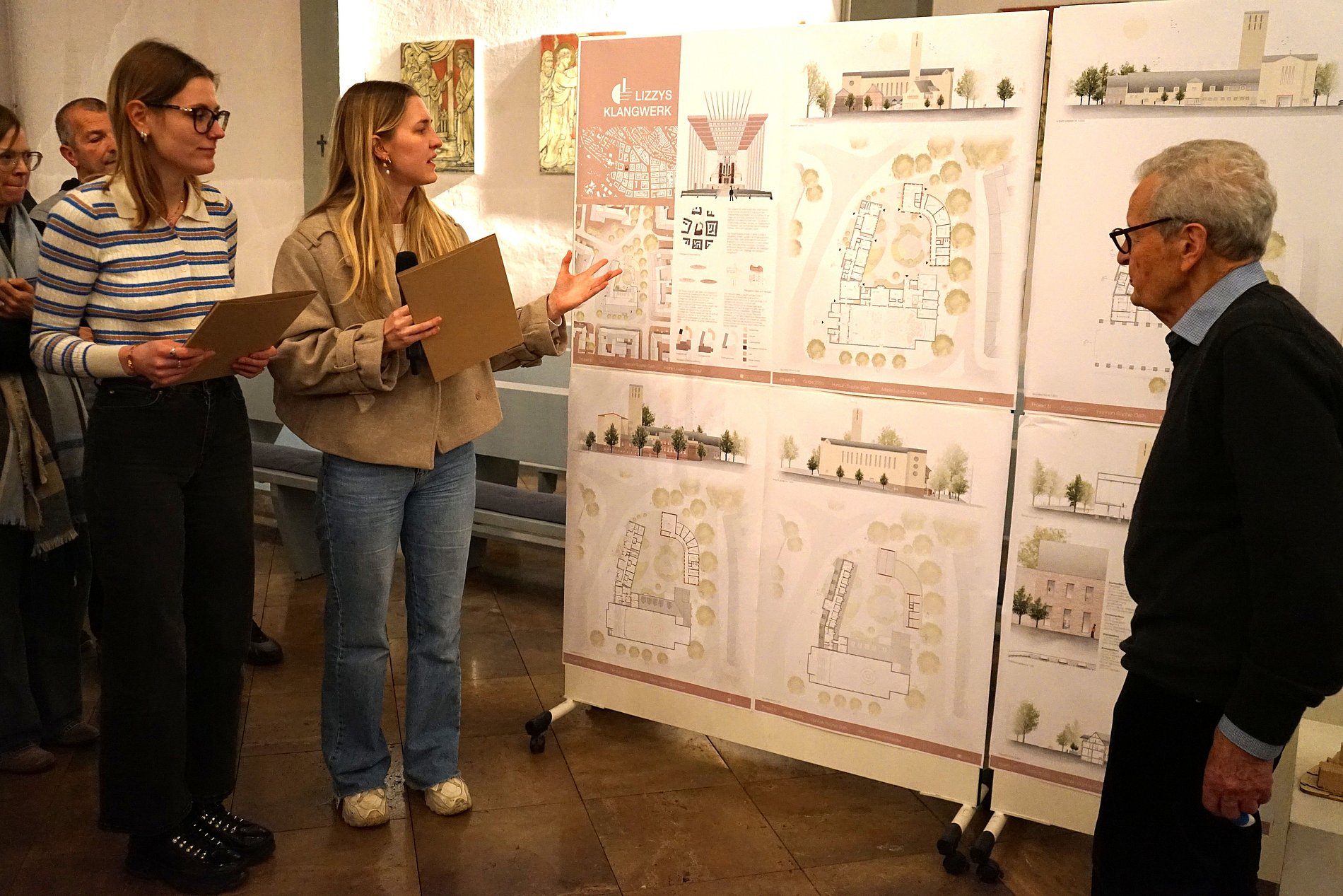 Die beiden Architekturstudentinnen stehen links neben ihren Planungsskizzen und erklären den Inhalt. Das Jurymitglied Peter Lesko steht rechts im Bild und hört zu.