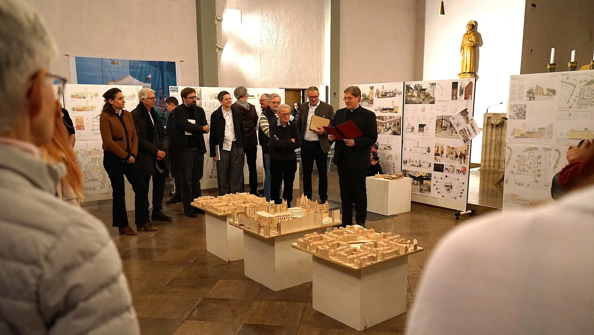 Pfarrer Klaus Nebel hält ein Mikrofon in der Hand und spricht zu den Anwesenden, die sich um die Modelle aufgestellt haben.