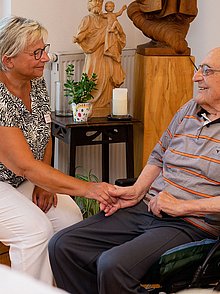 Eine Frau reicht einem älteren Mann im Rollstuhl die Hand. Sie ist für ihn da.